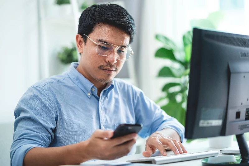 A man wearing glasses is sitting at a desk with phone in his hand